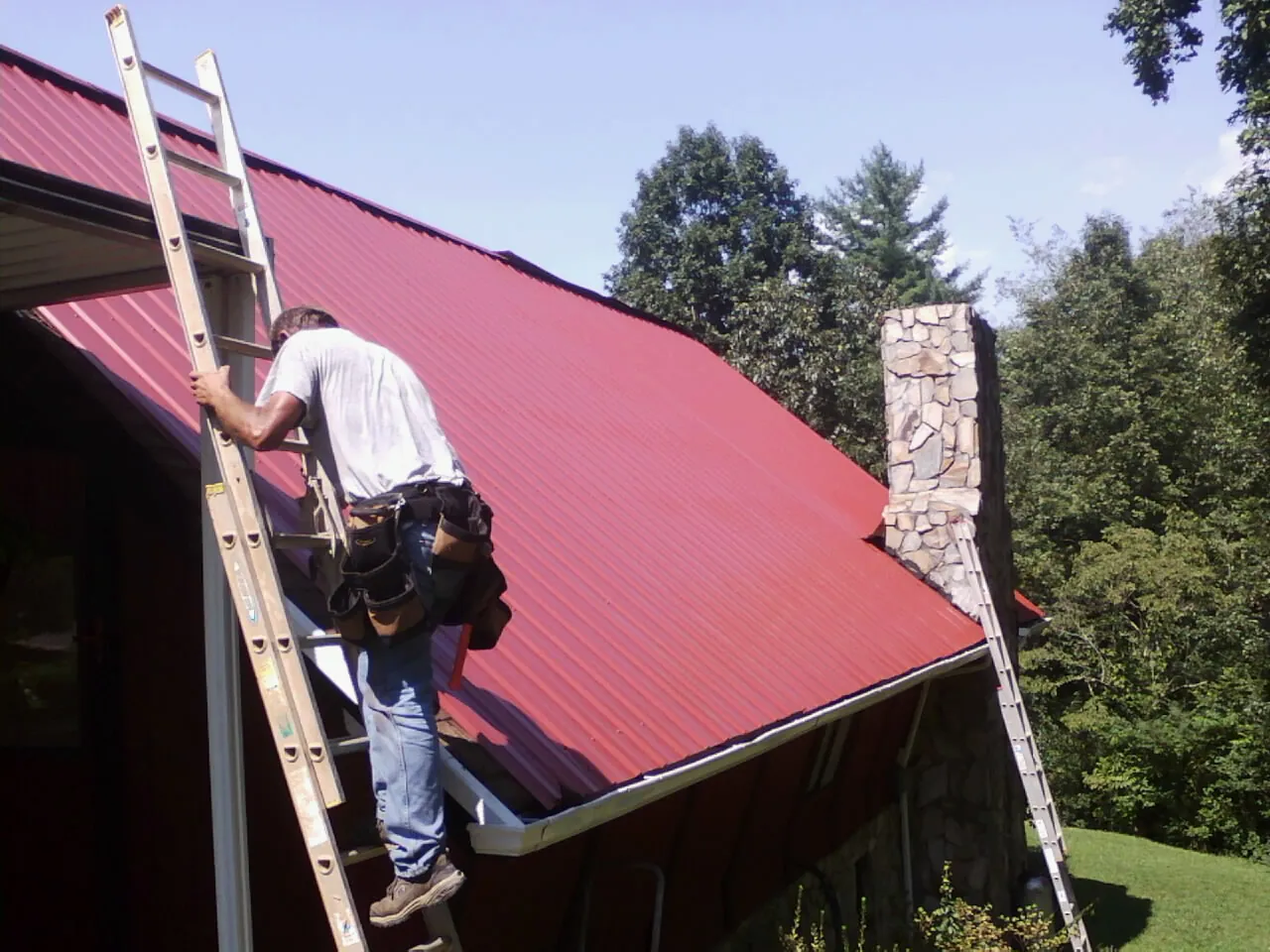 Roofer on a ladder beside a red metal roof and stone chimney — Soffit & Fascia Repair by Roofing Brothers in Chimayo